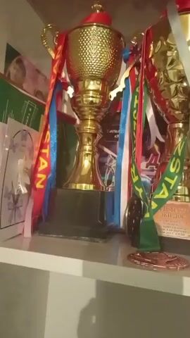 A close-up video shows several trophies and medals displayed on a shelf. The trophies are golden with various colored ribbons and some have inscriptions. There are also at least two medals visible, one with a green ribbon and another with a red and blue ribbon. The background is indistinct but appears to be a wall with some framed pictures or certificates. The lighting is dim and appears to be artificial. The shelf is white and there is some shadow cast on it. The video is shaky and out of focus in places.