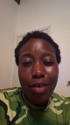 A close-up video shows a young Black person with short, dark hair, wearing a green camouflage shirt. The person looks directly at the camera and speaks. The background appears to be a room with a light-colored wall and a door. The lighting is bright, illuminating the person's face.