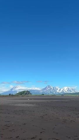 The video opens with a panoramic view of a black sand beach under a clear blue sky. In the distance, snow-capped mountains rise majestically, some with wisps of clouds clinging to their peaks. Several vehicles, possibly off-road SUVs, are scattered across the beach and the land in front of the mountains. The scene transitions to a closer view of the beach and then pans to the left, revealing rolling green hills that slope down towards the water. The waves are gentle, lapping against the shore. The video continues to pan, showing more of the green, tree-covered hillsides.