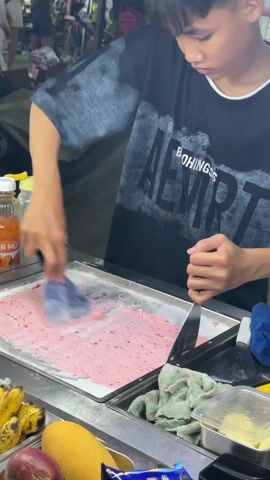 A young man is making rolled ice cream on a stainless steel cold plate in an outdoor market. He is using two metal spatulas to spread and scrape a pink mixture across the plate. There are bananas, a mango, and other food items on the counter near the cold plate. Bottles of juice or syrup are visible. The man is wearing a black t-shirt. Other people are visible in the background.