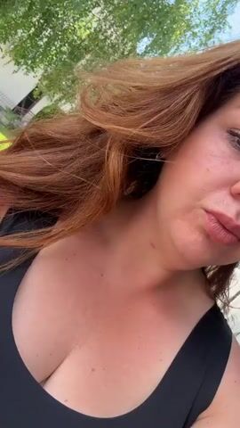 A woman with brown hair and a black tank top is taking a selfie outdoors on a sunny day. She slightly shifts the camera angle, showing a residential area with green lawns and houses in the background. There are trees visible, and a white truck is parked in the distance. The woman has a slightly smiling expression on her face.