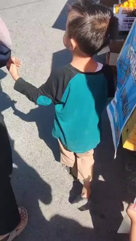 A child wearing a black and teal shirt and tan pants is walking away from the camera, holding hands with an adult whose arm is visible on the left. The adult is wearing a dark top and appears to be holding a plastic bag. The child's hair is short and dark, and they are wearing dark shoes. They are on a paved surface, possibly a street or marketplace, with shadows indicating it's daytime. In the background, there are boxes and what appear to be oranges on display, suggesting a market stall. Another adult, wearing a light pink headscarf, is partially visible on the right, looking down and holding a plastic bag. The scene suggests a casual outing or shopping trip.