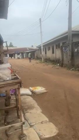 The video shows a dusty street scene in what appears to be a small town or village. The road is unpaved and reddish-brown. On the left side of the frame, there is a small wooden stand in front of a shop that appears to sell various goods. On the stand, there is a clear plastic container with what looks like balls of food inside. Bags are stacked on the ground nearby. A person wearing a patterned shirt and carrying a dark bag is walking away down the road. Buildings line both sides of the street. The sky is overcast.
