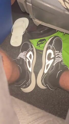 A close-up shot shows a seated person's legs and feet. The person is wearing black and white sneakers with thick white soles and white shoelaces. A pair of white flip-flops and a green bag are nearby on the carpeted floor of what appears to be a vehicle, possibly a bus or airplane. The person's hairy legs are visible, and they are wearing black socks. The background includes part of a blue object and a seat.