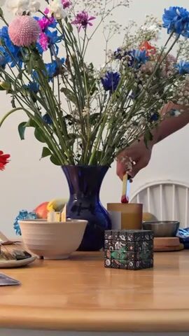 A person's hand is seen holding a stick of palo santo above a lit candle. The palo santo stick is being touched to the flame of the candle. The palo santo stick is lit, and a small flame is visible at the tip. The candle is in an amber-colored glass jar. In the background, there is a blue vase filled with flowers. To the left of the candle is a white bowl and a small cube with a space-themed pattern. The scene appears to be set on a wooden table.