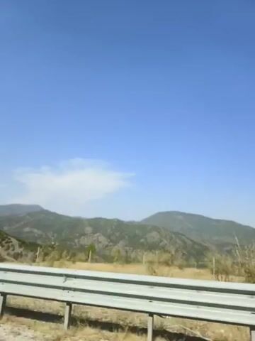 The video shows a landscape of mountains and a blue sky. A guardrail runs across the bottom of the frame. The mountains appear to be covered in greenery and rocks, and there is a small cloud in the sky. The scene is well-lit, suggesting it is daytime.