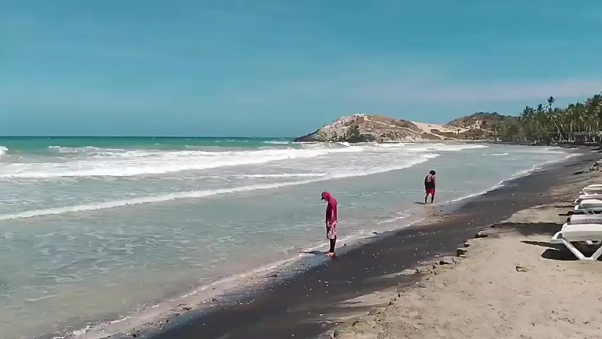 The scene shows a beach with a dark strip of sand along the water's edge, contrasting with the lighter sand further up the beach. Two individuals are visible: one stands at the water's edge in a red hooded shirt and dark shorts, looking down at the water, while the other walks further down the beach in a black shirt and red shorts.  The ocean has moderate waves breaking along the shore. In the background, there's a hill or small mountain, and palm trees are scattered along the edge of the beach.  Several white lounge chairs are neatly arranged on the sand near a structure with glass windows, possibly a beachside bar or restaurant. The sky is clear and blue.