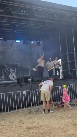 The video shows a band performing on a stage outdoors. The band consists of a drummer, a vocalist, a saxophone player, and a guitarist. In front of the stage, there is a barrier, and beyond that, several people are visible. A man wearing a light-colored t-shirt and shorts is bending down, appearing to be interacting with something on the ground. Two young children are also in the foreground; one is wearing a pink dress and the other is lying on the ground. A woman on the left is holding a smartphone and appears to be recording the performance. The ground is covered in dry grass or dirt, and the sky is blue with some clouds.
