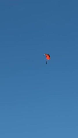 A skydiver is descending against a clear blue sky. The skydiver is wearing dark clothing and has an orange parachute deployed above them. The parachute appears to be stable and the skydiver is positioned directly beneath it. The skydiver is in the upper right quadrant of the frame. The video appears to be shot from below, looking up at the skydiver. The skydiver is slowly descending.