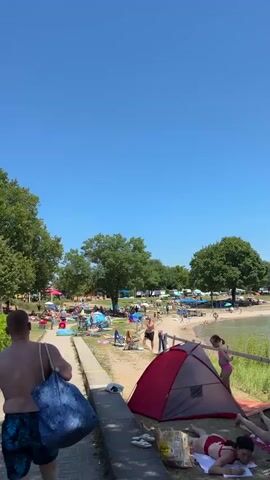 The video opens with a medium shot of a man walking away from the camera, carrying a large blue bag over his shoulder. He is wearing patterned swim trunks and is shirtless, revealing his back. The man is walking on a paved walkway bordered by a low concrete barrier on his right and a hedge on his left. Behind the hedge, a green leafy shrubbery is visible.

The walkway leads towards an open, sun-drenched area filled with people. In the background, there are numerous tents, umbrellas, beach chairs, and people scattered across a sandy and grassy area. Some individuals are lying on towels, while others are seated or standing. Trees are interspersed throughout the scene, providing shade in some areas.

To the right of the walkway, closer to the foreground, a red and grey tent is pitched on the sandy ground. Next to it, a person in a pink swimsuit is visible in the distance, standing near the edge of what appears to be a body of water. The sky above is a clear, vibrant blue, suggesting a sunny day.

As the video progresses, the camera pans slightly to the right and continues to follow the man. The background reveals more of the crowded beach area, with more tents, umbrellas, and people engaged in various activities. In the far background, there are more trees and some structures, possibly buildings or a campsite.

The camera then shifts its focus to the general scene, showing a wider view of the beach. People are seen relaxing, playing, and enjoying the water. The scene is lively and bustling with activity.

Later in the video, the perspective shifts again, focusing on a section of the beach with several people. A woman is sitting on a towel, and a man is standing nearby. Further in the background, a German flag is visible flying from a pole in front of a building. The overall impression is of a crowded public beach or recreational area on a warm, sunny day.