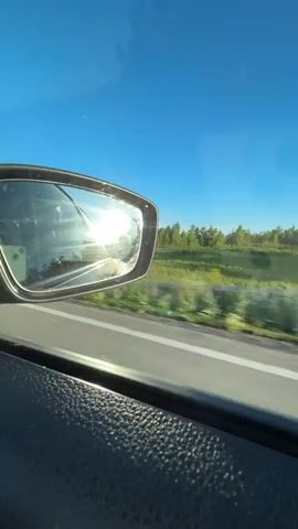 The video shows a view from inside a moving vehicle. The camera is positioned to capture the side mirror, which reflects the bright sun and the road behind the car. To the right of the mirror, the landscape is visible: a green grassy field bordering a tree line against a clear blue sky. The car appears to be traveling on a paved road, indicated by the white line on the right side. The interior of the car is also partially visible, showing a textured surface on the door or dashboard in the foreground. The sun is casting a glare, making the reflection in the mirror bright and somewhat indistinct.