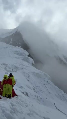 A group of people in brightly colored winter gear stand near several tents set up on a snowy mountain slope. The environment is rugged and mountainous, with large snow formations and peaks visible in the background, partially obscured by clouds. The tents are red and appear to be designed for extreme weather conditions, with one tent displaying the logo "PIONEER CAMP". One person is adjusting their jacket, while others are standing and observing the surroundings. The overall scene conveys a sense of expedition or climbing activity in a challenging, high-altitude environment.