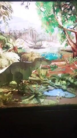 This is a close-up, medium-angle shot of a diorama depicting a prehistoric jungle scene. The diorama features several dinosaur figures, including a large, light green Tyrannosaurus Rex in the foreground, partially obscured by lush green foliage. Behind it, another dinosaur figure, possibly a Triceratops, is visible. The background is a painted mural of a dense jungle with a body of water. The diorama is lit from above, casting some shadows and highlighting the vibrant colors of the plants and dinosaurs. The scene appears to be static, with no discernible movement or people present. The overall impression is of a detailed and colorful model of a dinosaur habitat.