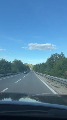 The video shows a view from the inside of a car traveling on a two-lane highway. The road is paved and has a white line marking the lanes. On the left side of the road, there are trees and bushes, and on the right side, there is a guardrail and more trees. The sky is blue with a few white clouds. There are several cars visible on the road ahead, as well as a motorcycle. The camera is pointed forward, showing the road stretching into the distance. The reflection of the car's interior is visible on the lower part of the screen. The time stamps indicate the video is a short loop.