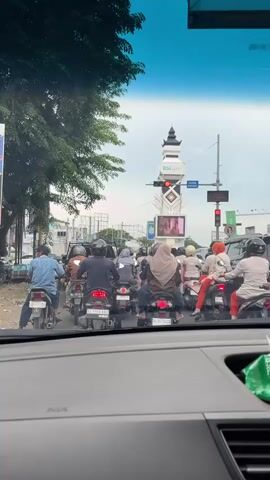 A traffic jam is depicted from the perspective of a car. Multiple motorcycles are lined up on the road, with riders facing away from the camera. In the background, a clock tower with a pointed roof stands tall. Traffic lights are visible, showing red. The sky is overcast.