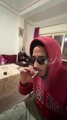 Here's a description of the video:
A young man wearing a red hoodie with the words "JAPAN RAGS CHICAGO" printed on it and sunglasses is indoors, smoking a joint. He exhales a large cloud of smoke towards the camera. A table with a patterned tablecloth, a water bottle, and other small objects is visible in the background. There's also a cabinet with a fan on it and a sofa with a red cushion visible in the background. The room appears to be lit by natural light from a window. Electronic music plays in the background.