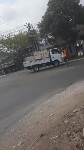 A person wearing an orange vest and dark shorts stands on the side of a road, facing away from the camera, with their arms extended to the sides. To the left of this person is a white pickup truck with a flatbed loaded with rectangular objects, possibly construction materials. The truck appears to be stopped. A silver SUV is driving past the pickup truck. The environment is a paved road with a dirt shoulder on the right side. Trees and buildings are visible in the background, along with utility poles and wires. The sky is overcast.