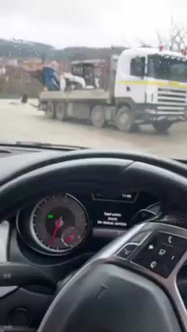 The video shows a view from inside a car, looking out at a rainy day. A large, white flatbed truck with a blue bobcat on it is parked on the side of the road. The car is driving on a wet road past the truck. A green van is seen in the distance. The dashboard of the car is visible, including the steering wheel, instrument cluster, and infotainment screen. The car appears to be driving slowly.