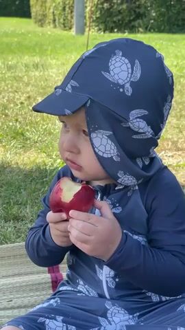 This close-up, medium shot captures a toddler sitting on a straw mat outdoors and eating a red apple. The child, wearing a dark blue swimsuit with a turtle pattern, is focused on the apple, with one hand holding it and the other near their mouth. The toddler has a protective sun hat, also adorned with a turtle print, that covers their head and neck. The background consists of a grassy lawn and a blurred fence, suggesting a sunny day. The lighting and the child's relaxed posture imply it's daytime, likely warm weather given the swimwear. The overall mood is peaceful and domestic, depicting a child enjoying a snack during an outdoor activity. There is no visible text in the image.