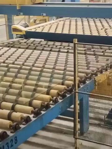 The video shows a conveyor belt system in an industrial setting. The conveyor belt is made up of many small rollers, which are moving objects forward. In the background, there is a larger conveyor belt with what appears to be tiles on it. The overall environment seems to be a factory or warehouse. There are no people visible in the video. The focus is on the machinery and the movement of objects along the conveyor belts.