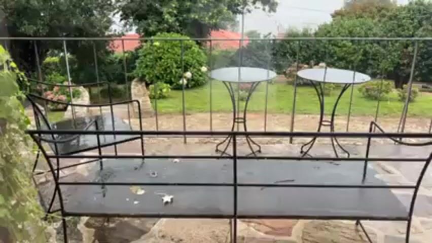 The video shows a rainy outdoor scene, focused on a stone patio furnished with black metal furniture. A bench and chairs are visible, along with two small round tables in the background. The patio is wet from the rain, and the surrounding garden is lush with greenery. Rain falls steadily, visible as droplets in the air and as it accumulates on the furniture. The scene is peaceful and depicts a rainy day in a garden setting.