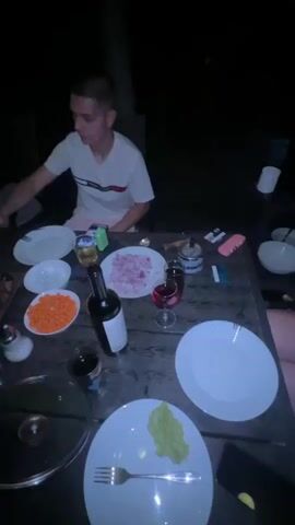 This video depicts an outdoor gathering at night, likely a dinner party, in Chaidefto, Greece. The scene is set at a table laden with food and drinks, suggesting a meal is in progress or about to commence. The lighting is dim, typical of an evening or nighttime setting.

Several people are present, though most are out of focus or partially visible. In the foreground, a person is seen holding a plate with food and a fork, indicating they are eating. Another person, in the middle ground, is seated and appears to be interacting with the food on the table. Towards the background, more individuals are visible, suggesting a group of friends or family enjoying each other's company.

The table is covered with various dishes, including what appears to be grilled meat, a bowl of chopped carrots, and a plate of chopped red onions. Bottles of wine and glasses are also present, along with plates, cutlery, and condiments. A bottle of what looks like sparkling water or clear spirits is also on the table.

The overall atmosphere appears relaxed and social. The focus is on the shared meal and the company. There is no discernible text visible in the video. The time of day is clearly night, and the location cues, while not overtly explicit, are consistent with a gathering in Greece, possibly at someone's home or a casual outdoor dining area.
