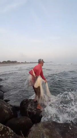 A man is standing barefoot on a large rock on a beach, preparing to cast a fishing net into the ocean. He is wearing a red long-sleeved shirt, brown shorts, and a baseball cap. He stands facing the ocean. The ocean waves break against the rocks as he prepares his net. The background shows a beach with buildings in the distance and a hazy, light-blue sky.