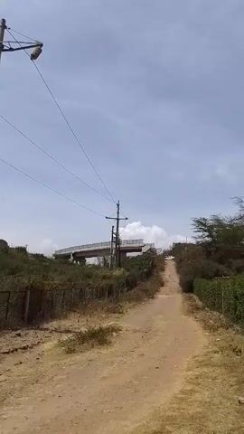 The video shows a dirt path leading up a hill toward a bridge. A chain-link fence lines the path on the left and right. On the left side of the path, there are two tall power poles with wires connected to them. The sky is light blue with scattered clouds. The vegetation appears dry and scrubby.
