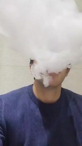 A person wearing a blue sweater exhales a large cloud of white smoke. The person's face is mostly obscured by the smoke, but parts of their mouth and chin are visible. The background is a plain, light-colored wall.
