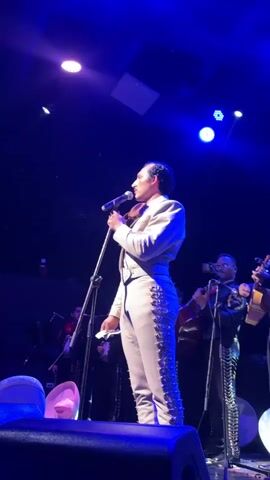 A mariachi band performs on a stage under blue lights. The lead singer, a man wearing a light-colored suit with ornate silver trim, holds a microphone and a violin. He is facing right, looking upwards and to the right. Behind him, other band members play instruments, including guitars and possibly violins. The stage has a dark background, and some seating or platforms are visible in the foreground. The lighting is dramatic, with spotlights creating highlights and shadows.