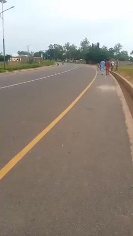 A video shows a paved road with a yellow line dividing the lanes. A boy in a grey shirt and yellow shorts is walking on the side of the road. In the distance, several people are walking together on the sidewalk. The environment appears to be rural with trees and some buildings visible. The sky is overcast.