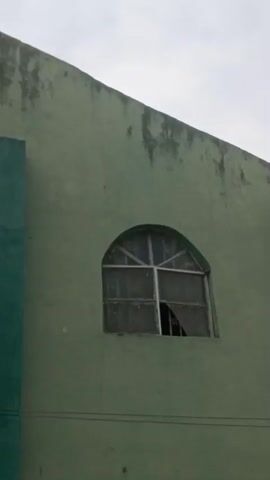 The video shows the upper portion of a pale green building. On the left side of the frame is a large emblem depicting a likeness of Jesus. The emblem is bordered by light blue and features text reading "THE SACRED CHERUBIM/SERAPHIM CHURCH, REF OF DEN UNIFICATION STREET PANLADA, OGUN STATE, NIGERIA." Below the Jesus image, it reads, "JESUS IS LORD." The right side of the frame features a vertical, rectangular column, also in light blue. Next to it, a window with a white frame has a broken pane. The overall texture of the building's exterior is uneven and slightly weathered. The sky is overcast. In the audio, voices and road traffic sounds are discernible.