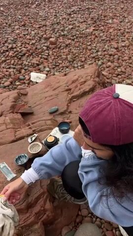 A person kneels on a large, red rock on a beach covered with red pebbles, setting up a tea service. The person wears a light blue sweatshirt, black leggings, and a burgundy and white baseball cap. A small tea set is arranged on a patterned cloth atop the rock, including a teapot, several small cups, and other tea accessories. The beach stretches to the horizon where the sea meets a grey sky. In the distance, a tall, rocky cliff rises from the water. The overall scene suggests a peaceful, contemplative moment in a natural setting.