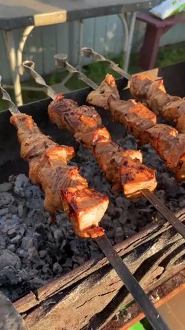 A close-up shot of several skewers of meat cooking over hot coals on a grill. The meat appears to be pork, cut into cubes and seasoned, with some pieces showing char marks from the grill. The skewers are arranged diagonally across the frame, with the closest one prominently featured in the foreground. The background is slightly blurred, showing a fence and some greenery, suggesting an outdoor setting. The lighting suggests it is daytime, possibly late afternoon due to the warm tone.