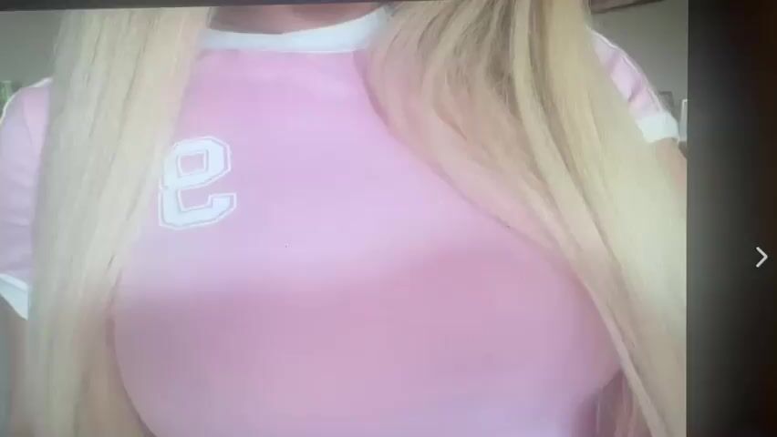 A close-up shot shows a person with blonde hair wearing a pink t-shirt with a white number "2" on it. The person's hands are visible in the frame, with one hand resting on their chest and the other touching their shirt. The background is blurred, but appears to be an indoor setting with some objects visible on a shelf. The lighting is bright and appears to be artificial.