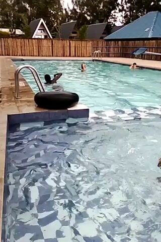 Here is a concise description of the video:

The video shows an outdoor swimming pool with several people swimming. In the foreground, a child wearing a yellow life ring is swimming with one arm extended. In the background, several other people are visible in the main pool, some swimming and others just standing in the water. A black flotation device is sitting on the pool edge near a set of steps with a railing. The pool is surrounded by a wooden fence and several small buildings with unique roof designs are visible behind it, along with some trees. The weather seems overcast. The water appears clear.