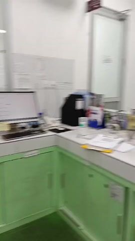 A person wearing a white lab coat and a white cap walks away from the camera into a laboratory. The lab has green cabinets and white countertops. There is a computer monitor on the counter. The camera pans to the right, showing more of the lab. There is a large cabinet with green accents and a window. The camera continues to pan, showing more lab equipment, including beakers and scales. There is a refrigerator and a microwave in the background. A clock is visible on the wall. There is also a fire extinguisher.