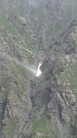 The video shows a mountainous landscape with gray rock and green vegetation. There is a small stream of water trickling down one part of the mountain. The mountain continues upwards to a peak with white snow. There are clouds in the sky.