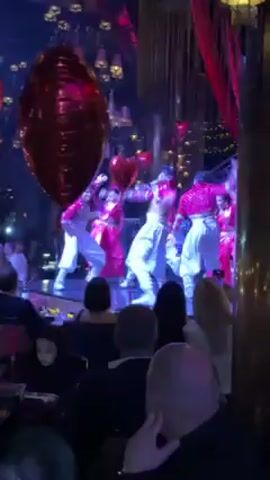 A group of dancers performs on a stage in front of a seated audience. The dancers wear white pants and matching shirts and jackets with pink accents. The performance is energetic with dancers leaping and spreading their arms, the video frame moves with their action. There are red heart-shaped balloons floating above the stage. The performance is taking place in a restaurant with dim lighting. The audience is watching the performance and appears to be enjoying it.