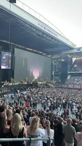 This media file captures a large outdoor concert at a stadium in Copenhagen, Denmark. The scene is dominated by a massive stage set up with elaborate, rocky structures and a giant screen displaying abstract, futuristic cityscapes in muted colors. The crowd, packed tightly into the stadium seating and standing areas, is a sea of faces illuminated by stage lights and the ambient daylight. Many people are looking towards the stage, some with hands raised, suggesting excitement and engagement with the performance.

The overall atmosphere appears to be one of anticipation and enjoyment. The time of day seems to be late afternoon or early evening, with a soft, diffused light indicating possibly overcast weather or approaching twilight. The stadium's architecture is visible in the background, including covered seating areas and supporting structures for the stage and sound systems.

A prominent piece of text is visible above the stadium seating: "VIER KØBENHAVN". This indicates the location as Copenhagen, Denmark, and "VIER" might be part of a sponsorship or stadium name. The crowd is diverse in age and attire, all focused on the event unfolding on stage. The sheer scale of the audience and the elaborate stage setup suggest a major musical performance or festival.