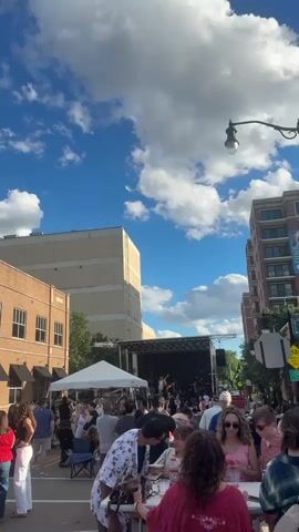This is a vertical, eye-level, full-shot video taken from behind a crowd of people looking at a stage setup on a street. The stage is located in the middle of a street, with a band playing. There are tents set up on the left side of the stage, and buildings line the street on both sides. The sky is blue with white, fluffy clouds. People are standing and sitting in front of the stage, some are holding drinks and looking at their phones. The overall atmosphere appears to be that of an outdoor event or festival.