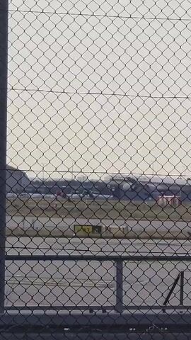 The video shows a chain-link fence in the foreground, with an airport runway and airplanes in the background. A sign on the fence reads "Achtung! No-Drone-Zone!". The sky is overcast.