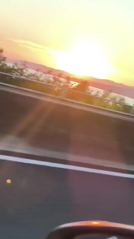 A sunset is visible over a body of water and distant hills. The sun is a bright orange orb in the sky, casting a warm glow over the landscape. The water reflects the colors of the sunset, creating a shimmering effect. Trees and vegetation line the foreground, with a road or highway visible in the lower part of the frame. The sky is a gradient of orange and pale blue, transitioning from the horizon to the top of the frame. The video appears to have been taken from a moving vehicle.