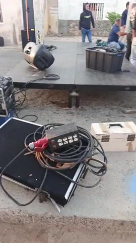 The video shows several people working on a makeshift stage in an outdoor setting. The stage is a raised platform with a metal frame and a black surface. Several individuals are on the stage, including one who appears to be adjusting something inside a black box. Another person is standing next to a large wooden panel. There is a speaker on the stage, and the area around it is littered with cables, a toolbox, and other equipment. Some of the individuals are wearing casual clothing and baseball caps. The background consists of houses and a clear sky. The overall impression is that the people are setting up or dismantling equipment for an event.