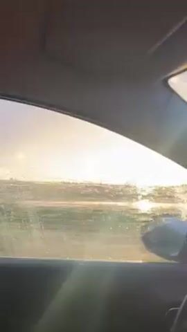 A car is driving on a wet road with green vegetation on both sides. The car's windshield wipers are on. The sky is overcast and there is a hint of light on the horizon. The car is following another vehicle.