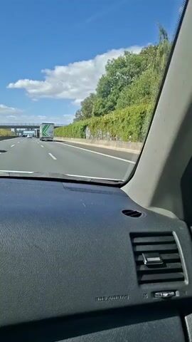 The video is taken from inside a car driving on a highway. The camera is positioned looking forward through the windshield. The dashboard of the car is visible in the foreground. Outside the car, a highway with multiple lanes stretches ahead. Several vehicles, including trucks and cars, are on the road. There is a green hedge lining the side of the highway, and trees are visible beyond the hedge. The sky is blue with white clouds, suggesting a sunny day. A highway sign structure is visible in the distance. The car appears to be moving at a moderate speed.