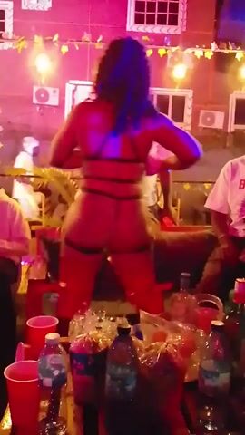 A woman with dark, curly hair dances on a table in an outdoor setting. She wears a revealing black lingerie-style outfit with multiple straps and high, red, glossy boots. The table is covered with drinks and snacks. Several people are seated or standing around the table, watching her.  The background includes a building with windows, air conditioning units, and string lights. The overall lighting is a mix of white and blue.