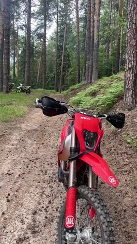 A static outdoor shot shows a red dirt bike on a dirt path, angled towards the viewer. The bike's front wheel, headlight, handlebars with handguards, and front fender are visible. The path is surrounded by tall trees and green foliage, with another dirt bike partially visible in the distance. The camera pans up to reveal more of the tall trees and green undergrowth.