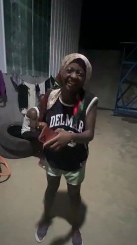 A medium shot shows a woman indoors holding two smartphones. She is light-skinned, wears a beige headscarf, a sleeveless black jersey with "DEL MAR 22" printed on it, and light-green shorts. She is smiling and looking directly at the camera. Behind her, a darker-skinned man is seated, also using a smartphone. In the background, there's a corrugated metal wall, clothes hanging on a line, and what appears to be a stack of tires. There is a short, metal structure behind the man.
