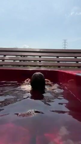 A woman emerges from a red-tinted pool on a sunny day. The woman has long, dark hair and is wearing a dark thong bathing suit. She places her hands on the side of the pool and lifts herself up, facing away from the camera. She then turns to look over her shoulder and splahs the water with her feet. A wooden bench is visible behind the pool, and a power line tower can be seen in the distance against a clear blue sky.